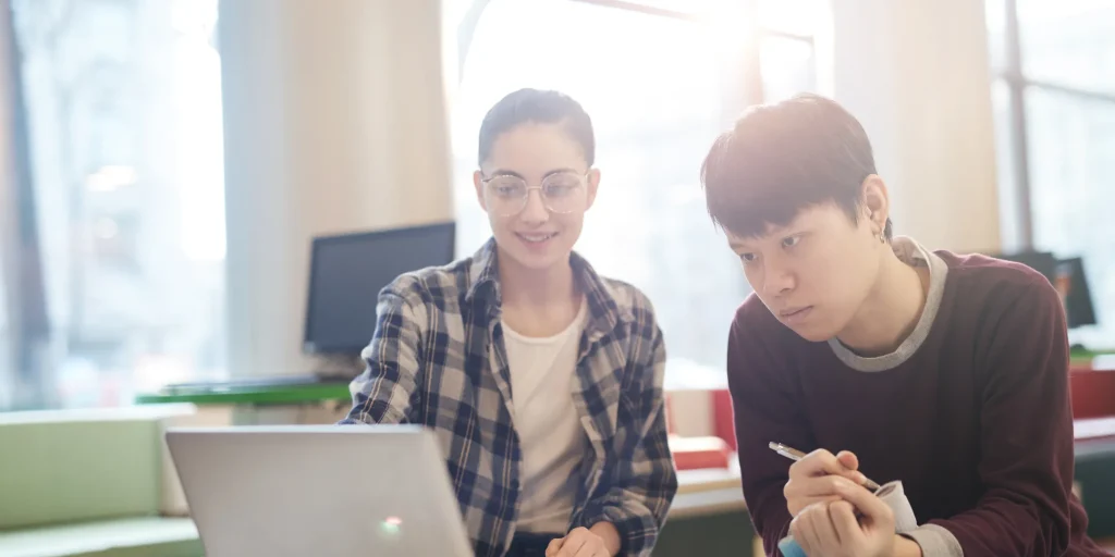 Two people sit side by side working on a laptop in a bright room. One smiles while pointing at the screen, and the other looks focused while holding a pen and notebook. Sunlight streams in from a window, creating a warm, studious atmosphere.