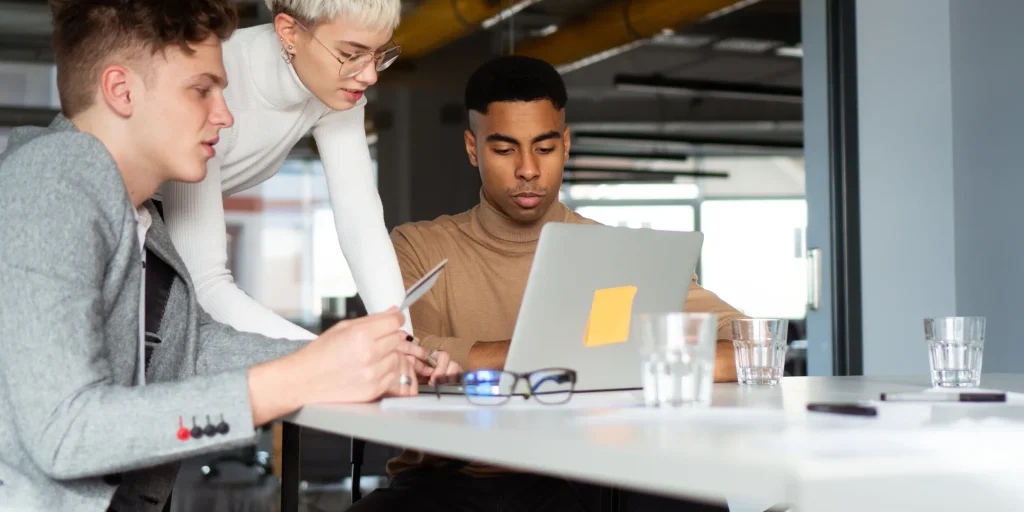Three coworkers gather around a laptop at a table, focused on the screen. One person points or holds a pencil while another leans in to look more closely. Glasses and water glasses sit on the table, and the setting suggests a modern office environment.