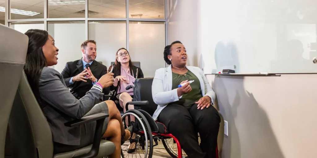 A group of colleagues sit together in a meeting room, engaged in discussion. One person using a wheelchair gestures toward a whiteboard while speaking, while others listen and respond. The setting appears professional and collaborative, with glass walls and office furniture in the background.