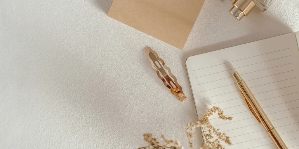A flat lay of minimal, neutral-toned stationery items arranged on a textured surface. A notebook with lined pages, a gold pen, a hair clip, a small spray bottle, and dried flowers create a calm, aesthetic composition.