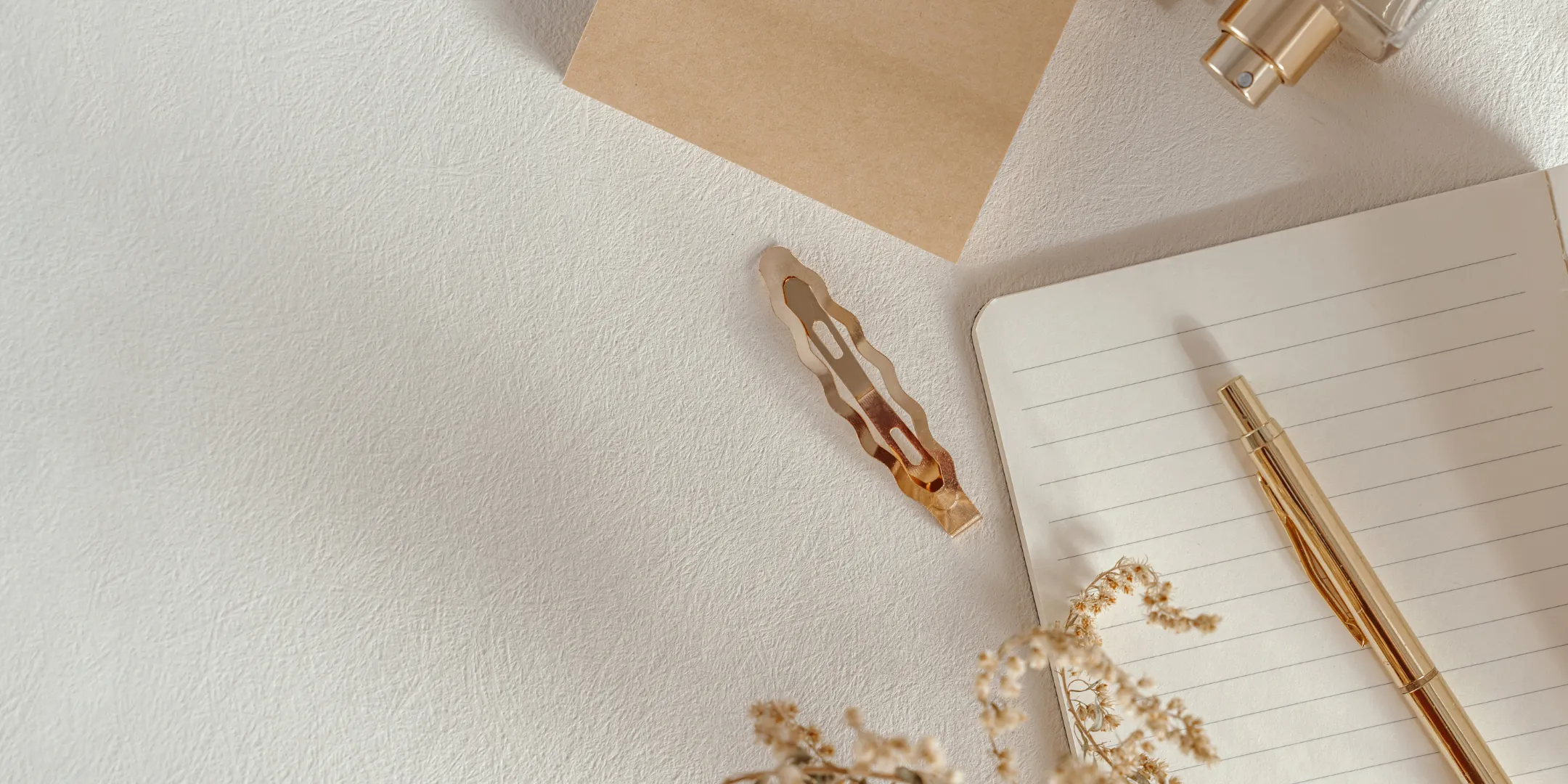 Minimal overhead workspace with an open lined notebook, gold pen, paper clip and neutral stationery arranged on a light textured surface.
