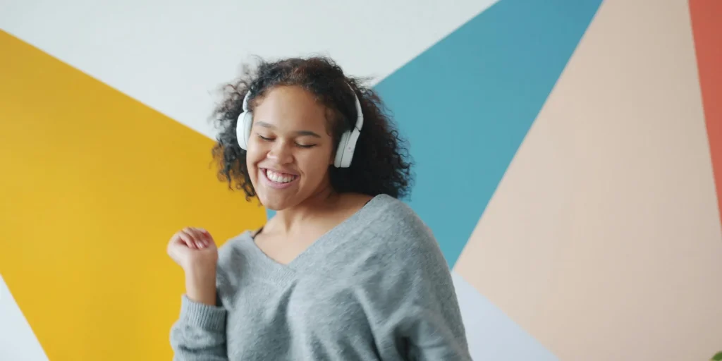A person wearing over-ear headphones smiles and moves as if dancing or enjoying music. They wear a casual grey sweater and stand in front of a colorful geometric background, creating a bright and energetic atmosphere.