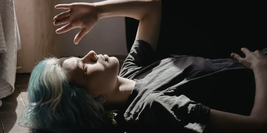 A person with short, light-colored hair lies on the floor near a window, partially illuminated by sunlight. Their arm is raised across their face, shielding their eyes from the light. The scene has a quiet, introspective mood, with strong contrast between light and shadow.