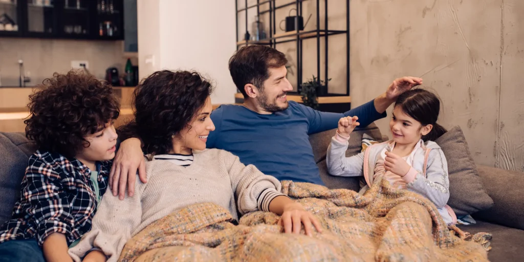 A family of four sits closely together on a couch in a cozy living room, sharing a relaxed and playful moment. A parent gently touches a child’s head while smiling, as the child gestures with their hand. Another parent and child sit nearby, watching and smiling. A soft blanket is draped over their laps, and the warm indoor setting creates a comfortable, affectionate atmosphere.