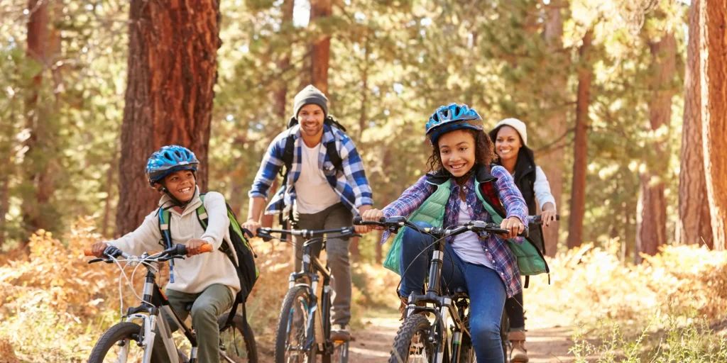 A family rides bicycles together along a forest trail, surrounded by tall trees and warm sunlight. Two children ride in front wearing helmets and backpacks, smiling as they pedal, while two adults follow closely behind. The scene feels active and joyful, with a sense of togetherness and outdoor adventure.