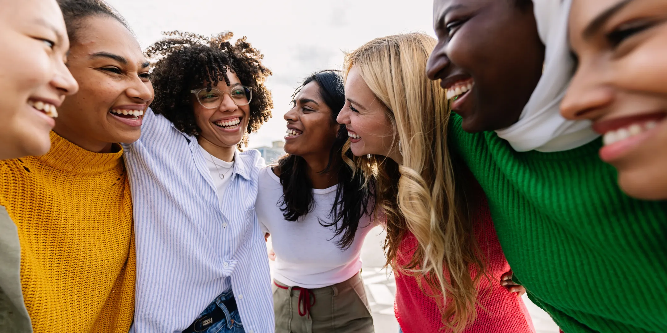 A diverse group of smiling young adults stand close together in a circle outdoors, laughing and looking at one another, conveying friendship, belonging, and connection.