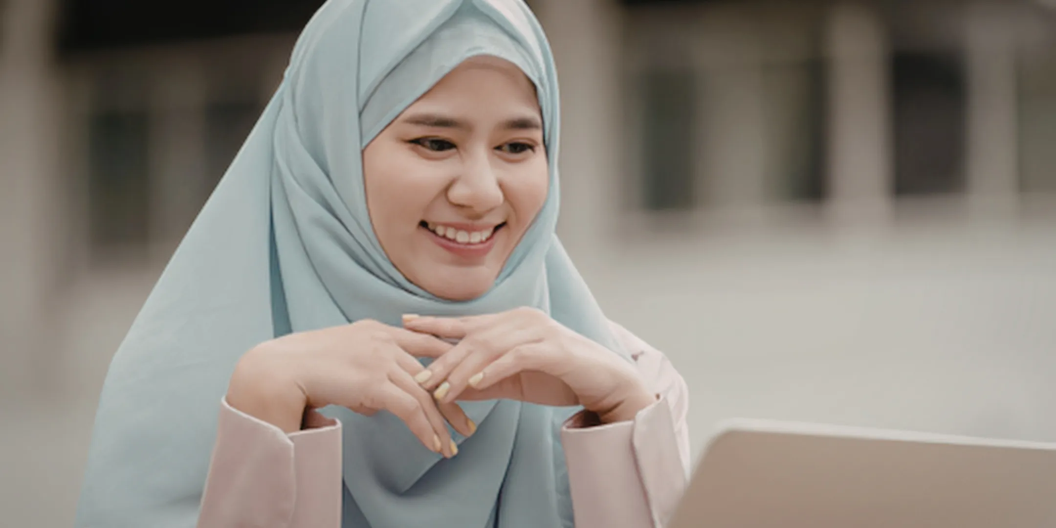 A person wearing a light blue hijab sits indoors, smiling gently while looking toward a laptop. Their hands are loosely clasped in front of them, suggesting they are engaged and attentive, possibly in a conversation or virtual meeting. The background is softly blurred, creating a calm and focused atmosphere.
