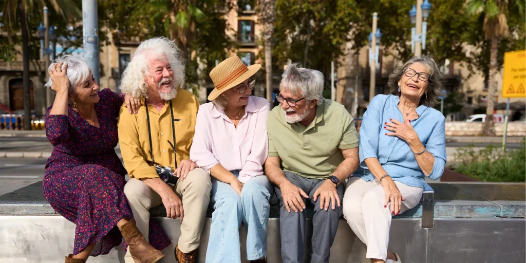 Five older adults sit together on a bench outdoors, laughing and enjoying each other’s company. They appear relaxed and connected, with some leaning toward one another and sharing conversation. Their clothing is casual and colorful, including a hat and a camera worn around one person’s neck. The setting includes palm trees and buildings in the background, suggesting a warm, urban outdoor space.