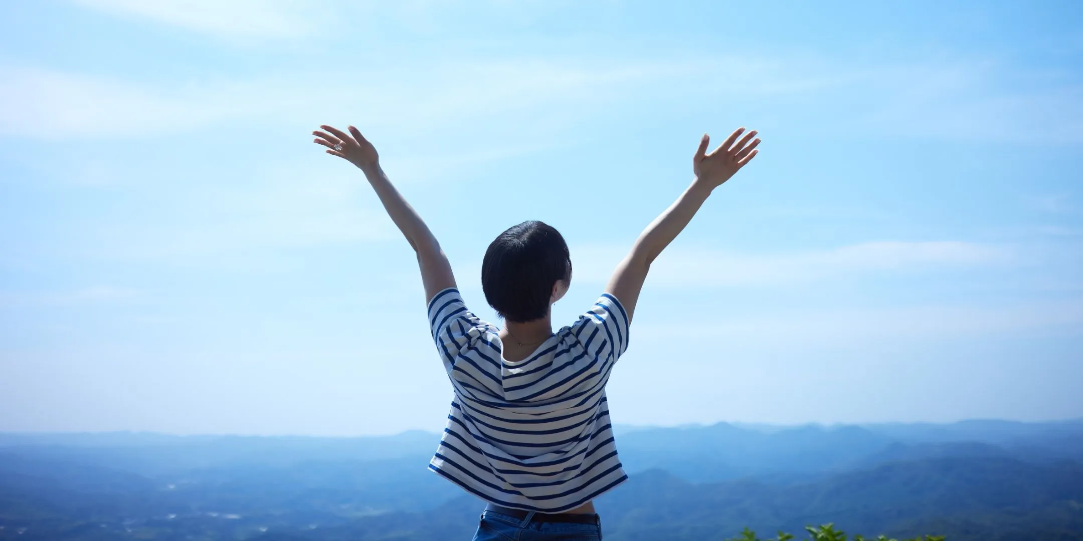 Person standing outdoors with their back to the camera, raising both arms toward a bright blue sky above a distant mountain landscape.