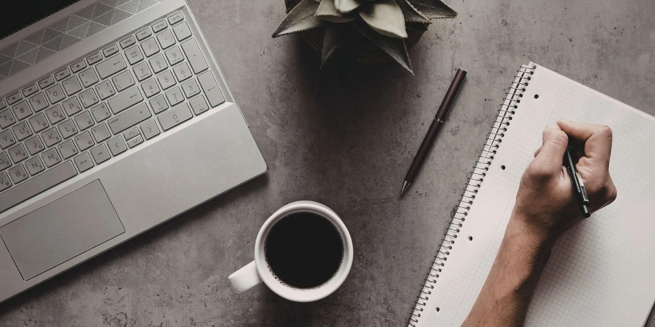 Overhead view of a workspace with a laptop, cup of coffee, notebook, pens and a small plant, while a person writes in the notebook.