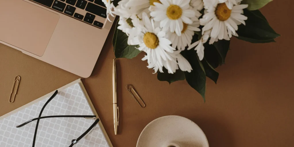 This image shows a neatly arranged workspace viewed from above, creating a calm and organised feel.  On the left, part of a laptop is visible, suggesting a work or study setting. Nearby, there’s a notebook with a subtle patterned cover, a pair of glasses resting on top, and a pen placed alongside it—items that hint at writing, planning, or reflection. A couple of paper clips are scattered lightly, adding to the everyday desk detail.  On the right, a bouquet of white daisies brings a soft, natural element into the scene, balancing the more structured objects with something gentle and grounding.  Overall, the image conveys a sense of quiet focus, organisation, and care—an environment that feels both productive and soothing.