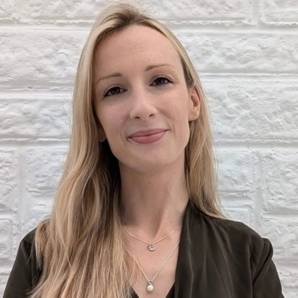 Smiling blonde person standing in front of a white brick wall.