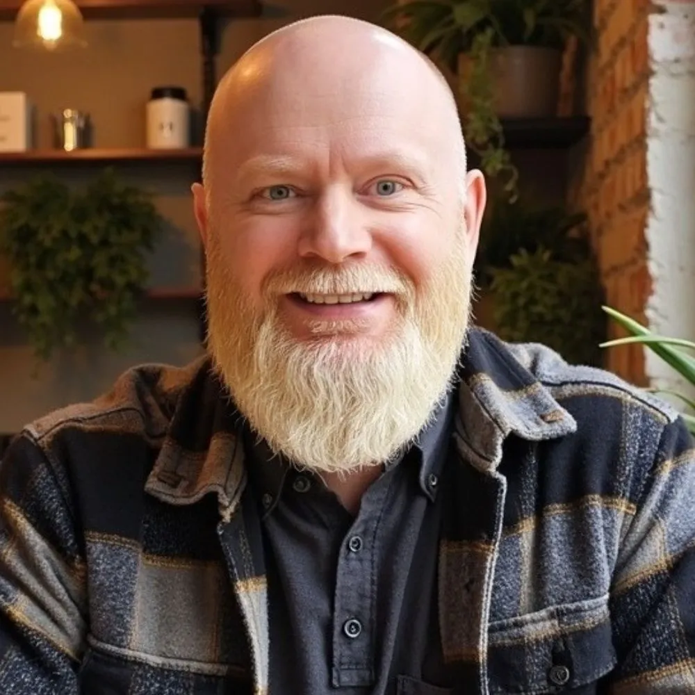 Smiling person with a white beard seated indoors with plants in the background.