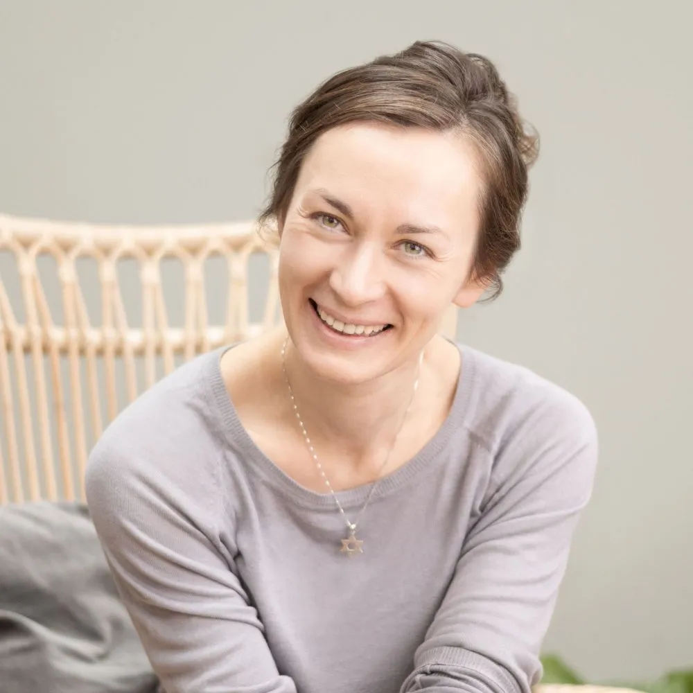 Smiling person with dark hair seated indoors against a neutral background.