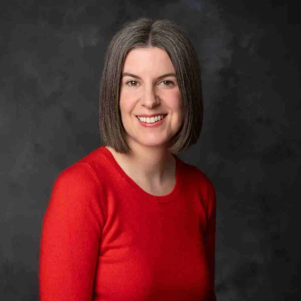 Smiling person with shoulder-length grey hair wearing a red top against a dark background.
