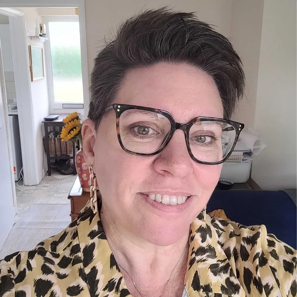 Smiling person with short dark hair, glasses, and patterned shirt taking an indoor selfie.