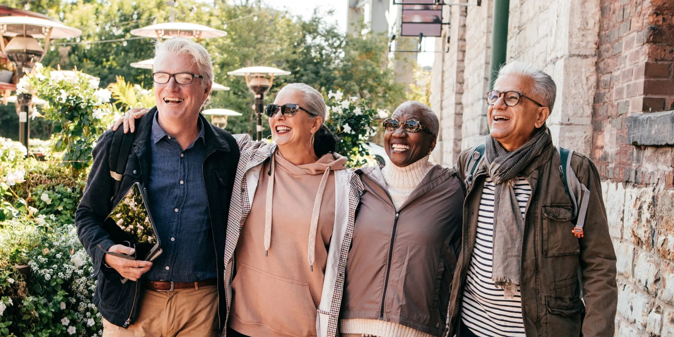 Diverse group of four older adults walking closely together outdoors, smiling and laughing in a sunny garden setting.