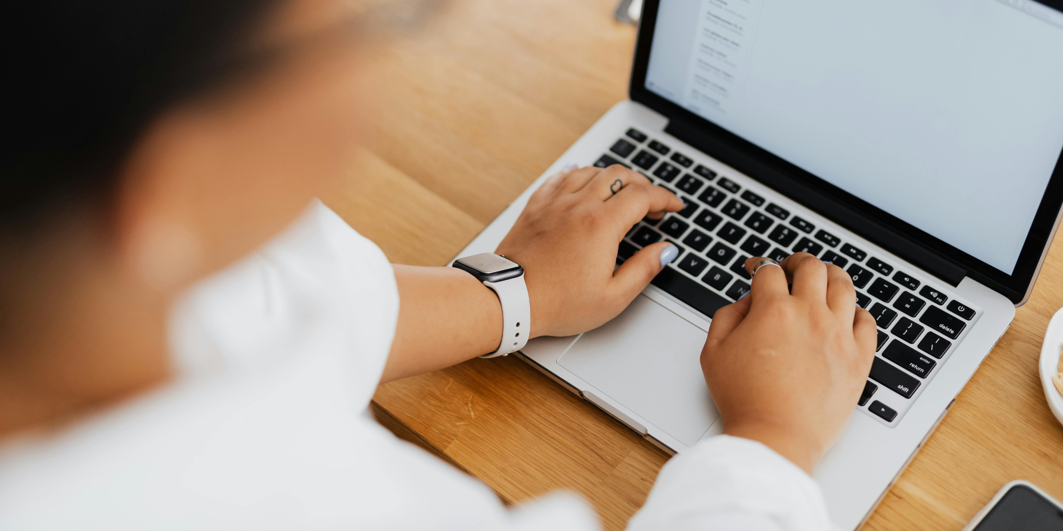 Close-up view of a person typing on a laptop at a wooden desk, with their hands on the keyboard. They are wearing a smartwatch and rings, suggesting focused work, study, or online communication in a modern workspace.