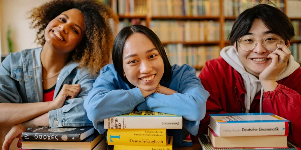 Three smiling young adults of diverse backgrounds lean on stacks of books in a library, looking toward the camera in a warm, welcoming study environment.