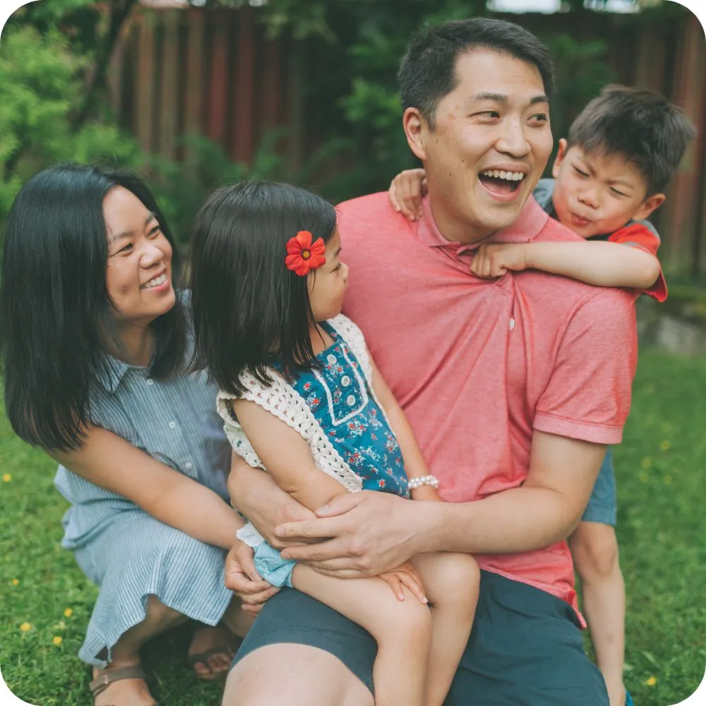 A smiling family of four enjoys time together outdoors in a garden. One parent holds a young child while the other child playfully hugs the second parent from behind, creating a warm and joyful family moment.