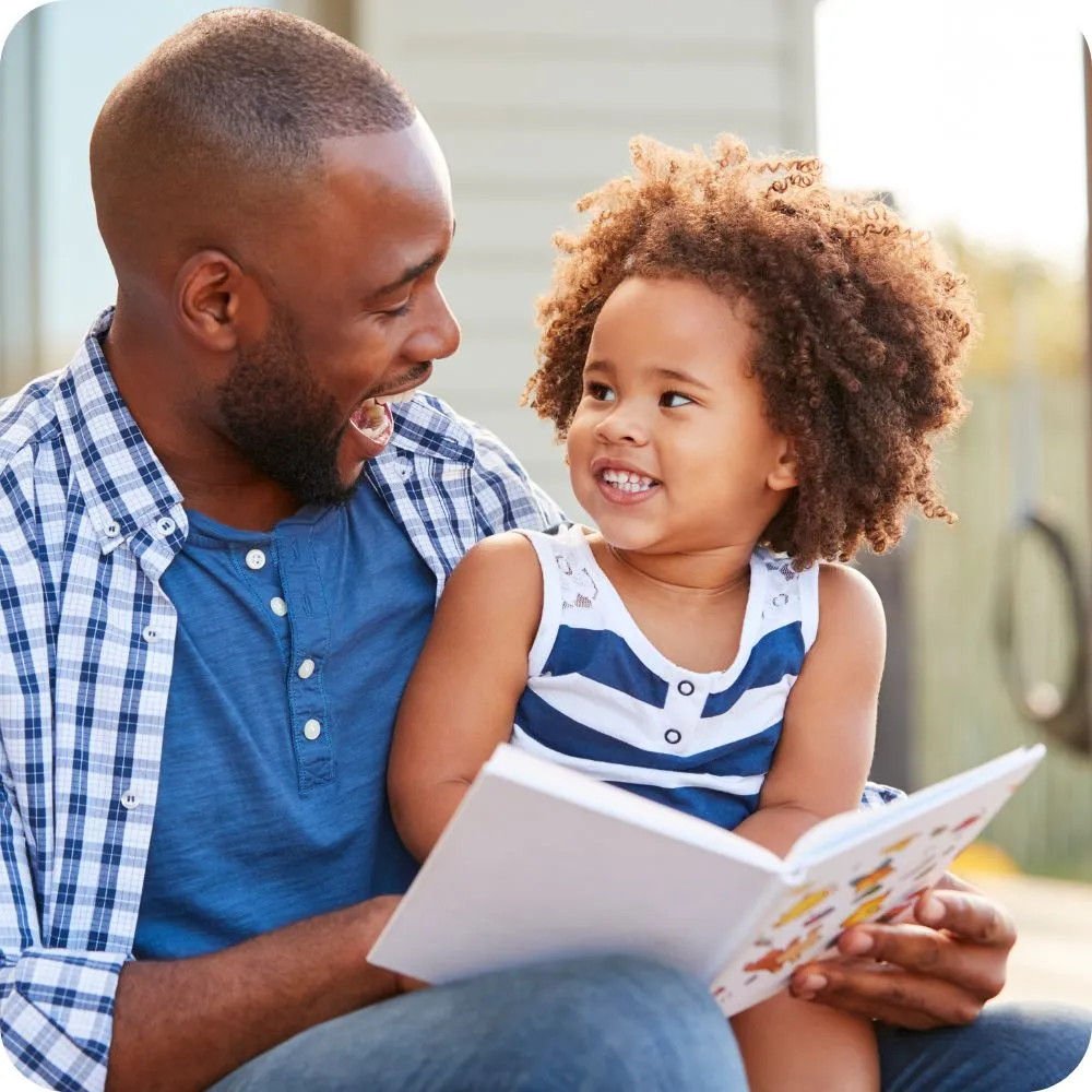 Father and child sit together reading a picture book. They smile at each other, sharing an engaging and affectionate moment during story time outdoors.