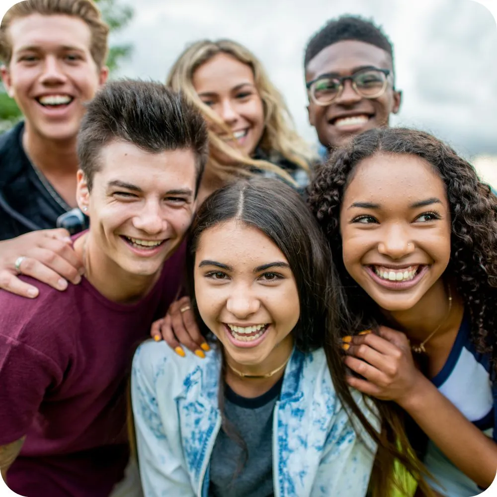 A diverse group of six smiling teenagers stand closely together outdoors, looking toward the camera. Their relaxed poses and cheerful expressions reflect friendship, belonging, and connection.