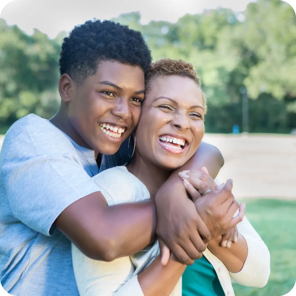 A smiling teenager hugs an older adult from behind in a park, with both laughing together. Their joyful expressions and close embrace convey warmth, affection, and a strong family bond.