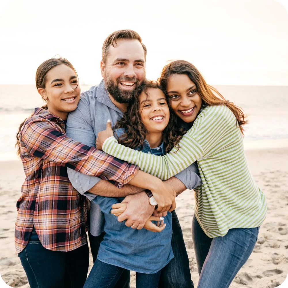 A family of four shares a group hug on a beach at sunset. The parents and two children smile warmly as they embrace, capturing a moment of love, togetherness, and happiness by the sea.