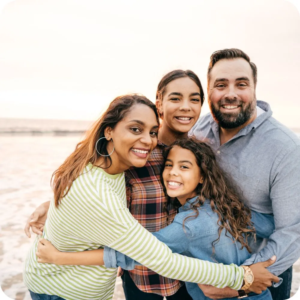 A smiling family of four embraces on a beach, posing together at sunset. 
