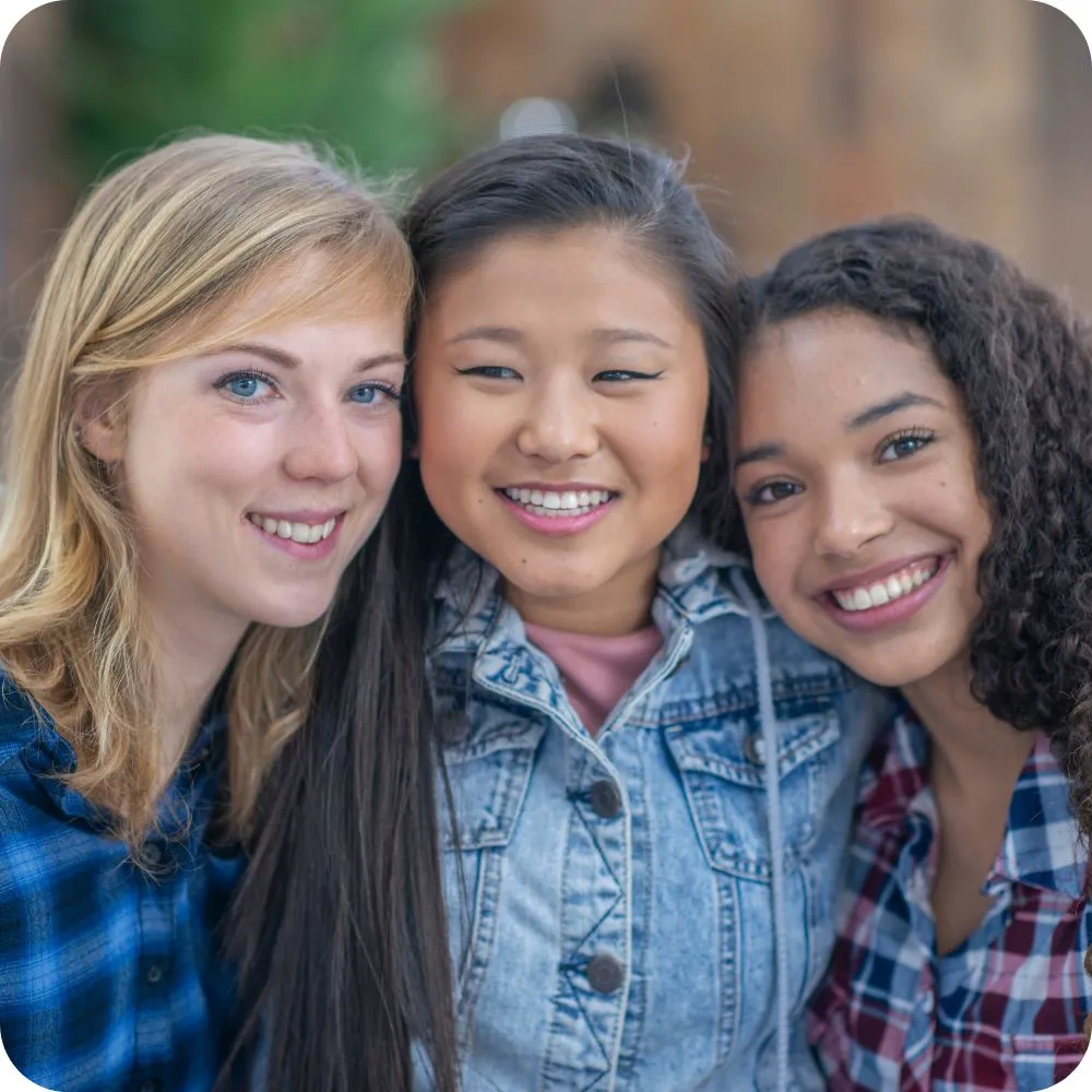 Three smiling teenagers from diverse backgrounds stand close together outdoors with their arms around one another.