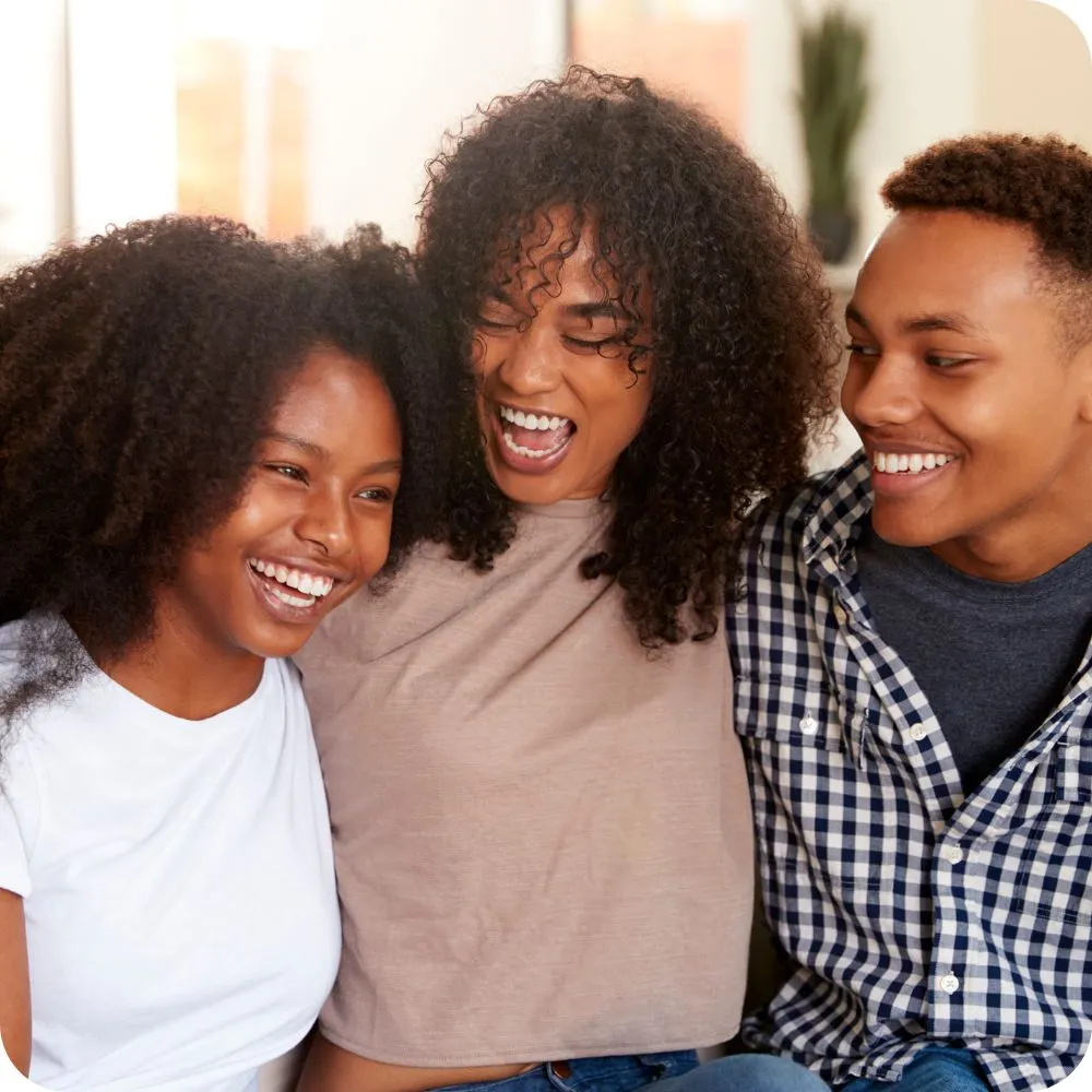 Mother and her 2 teenagers sit together indoors, laughing and enjoying each other’s company. 
