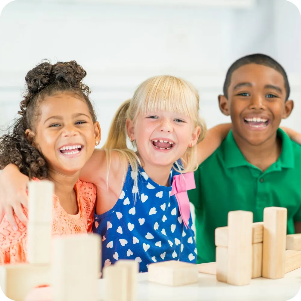 Three smiling children of diverse backgrounds sit side by side with their arms around each other, laughing together at a table with wooden building blocks in a bright classroom or playroom.