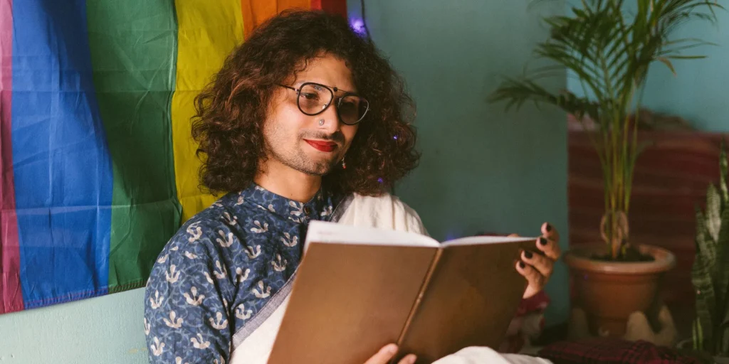 A person wearing glasses and reading a book sits indoors beside a rainbow Pride flag, conveying a calm, affirming, and welcoming environment.
