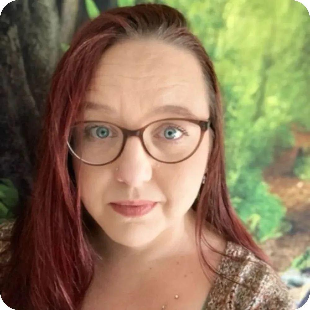 Person with long red hair and glasses standing in front of a leafy green background.