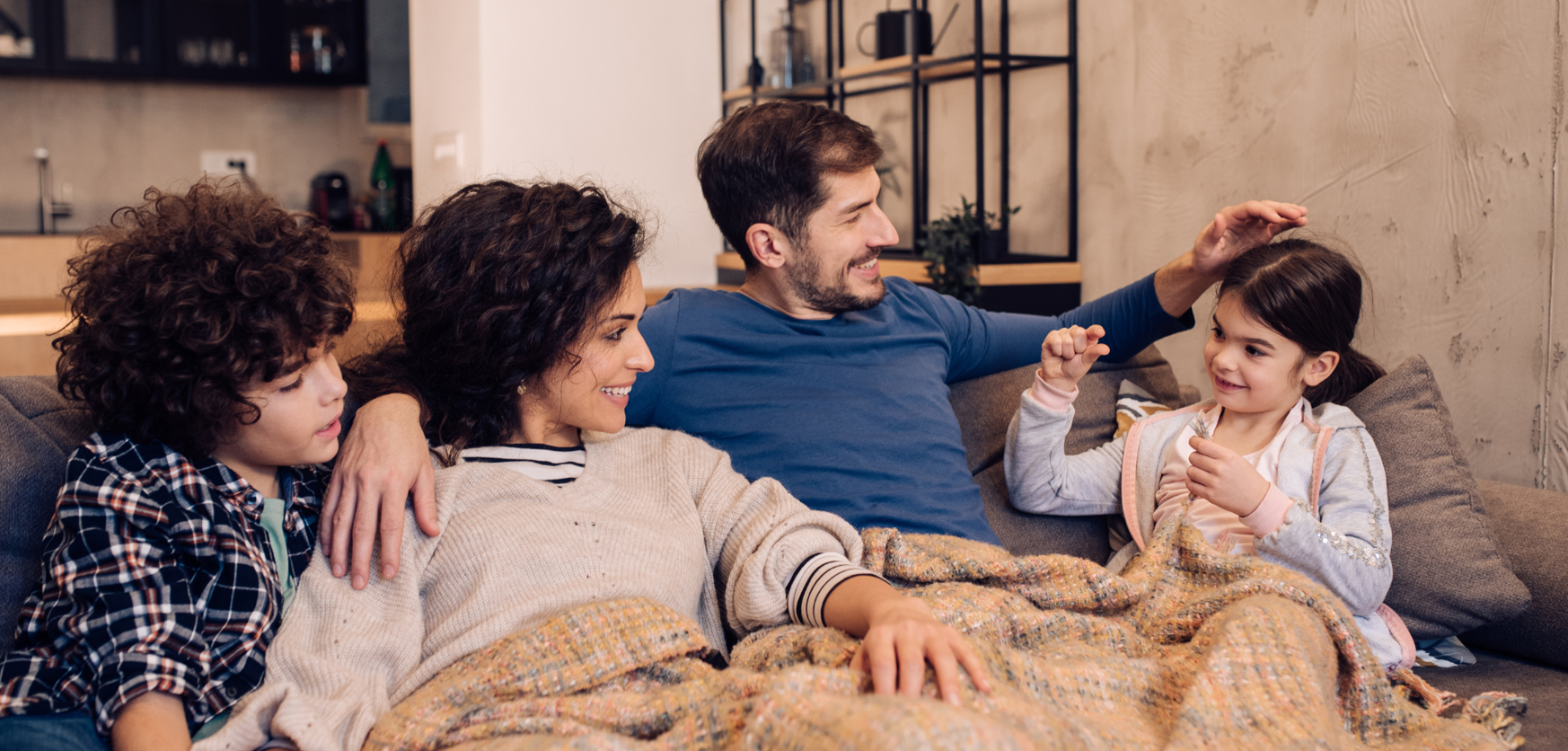 Family of four relaxing together on a sofa at home, smiling and interacting warmly under a shared blanket.