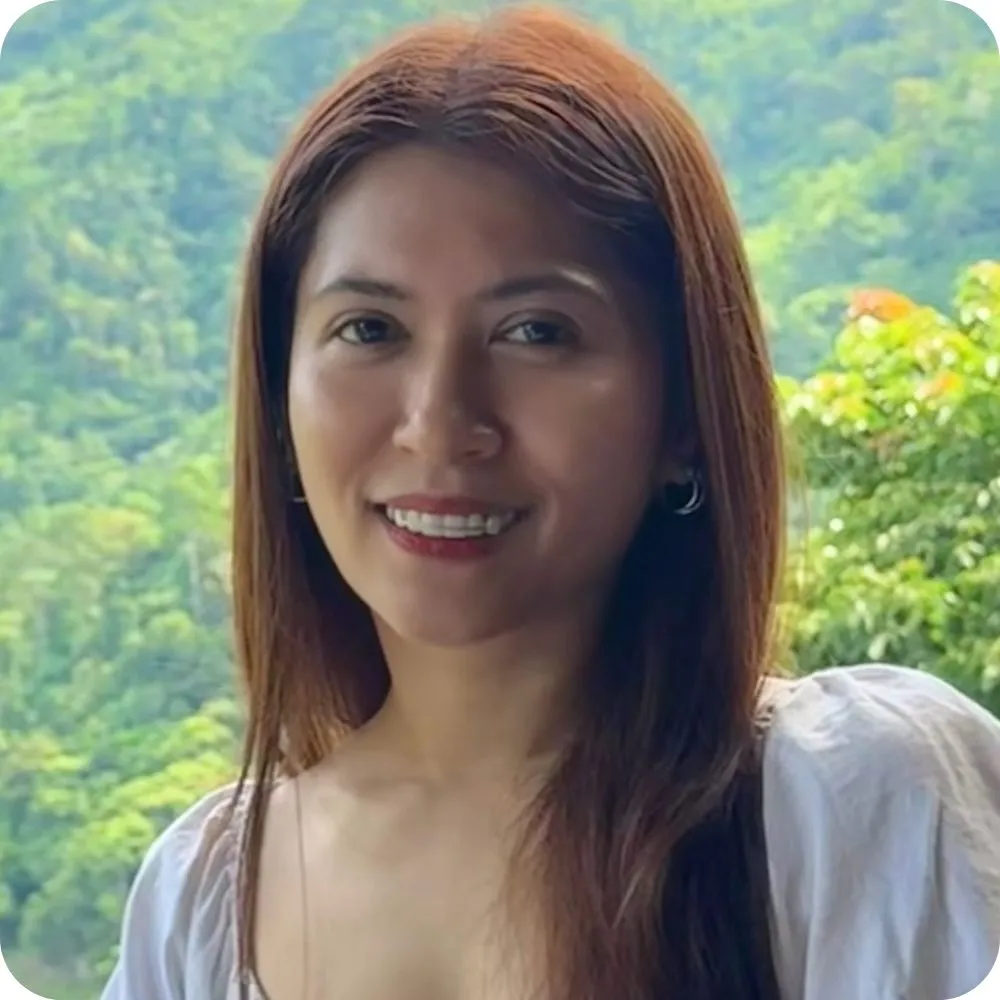 A smiling person with long dark hair wearing a white blouse stands outdoors with a lush green forest and hills in the background.