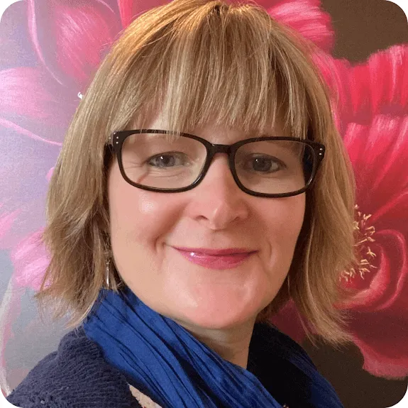 Smiling person with glasses, a blue scarf, and blonde bob haircut in front of a floral background.