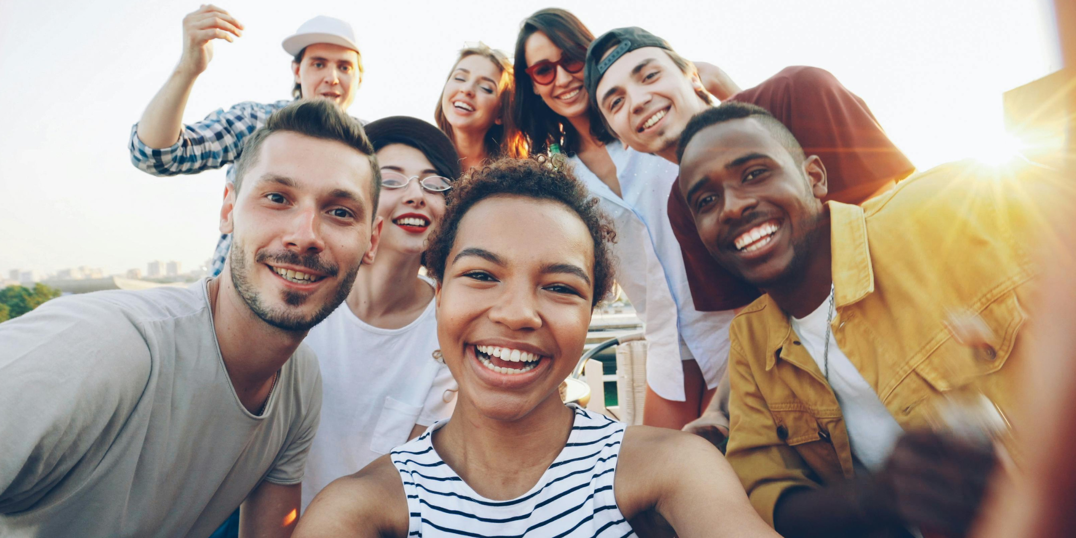 A diverse group of friends smile at the camera while taking a selfie outdoors, with sunlight in the background, conveying joy, friendship, and togetherness.