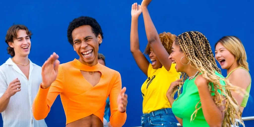 A diverse group of young adults laugh, celebrate, and dance together in front of a bright blue wall.