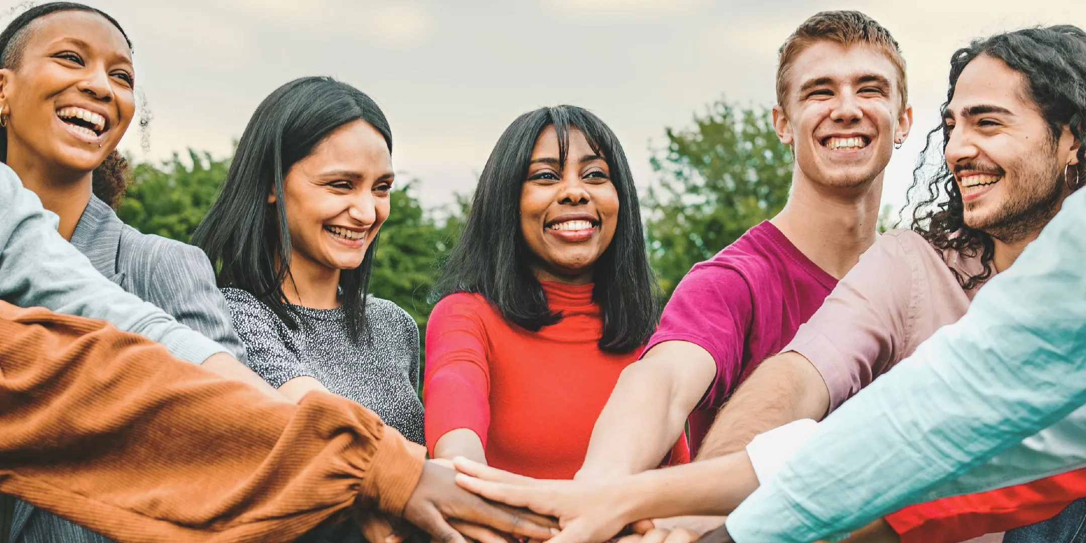 Diverse group of five adults smiling and laughing outdoors as they place their hands together in the centre, representing connection, community and teamwork.