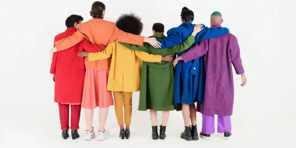 A diverse group of people stand arm in arm with their backs to the camera, wearing brightly coloured clothing to represent connection, belonging, and mutual support.