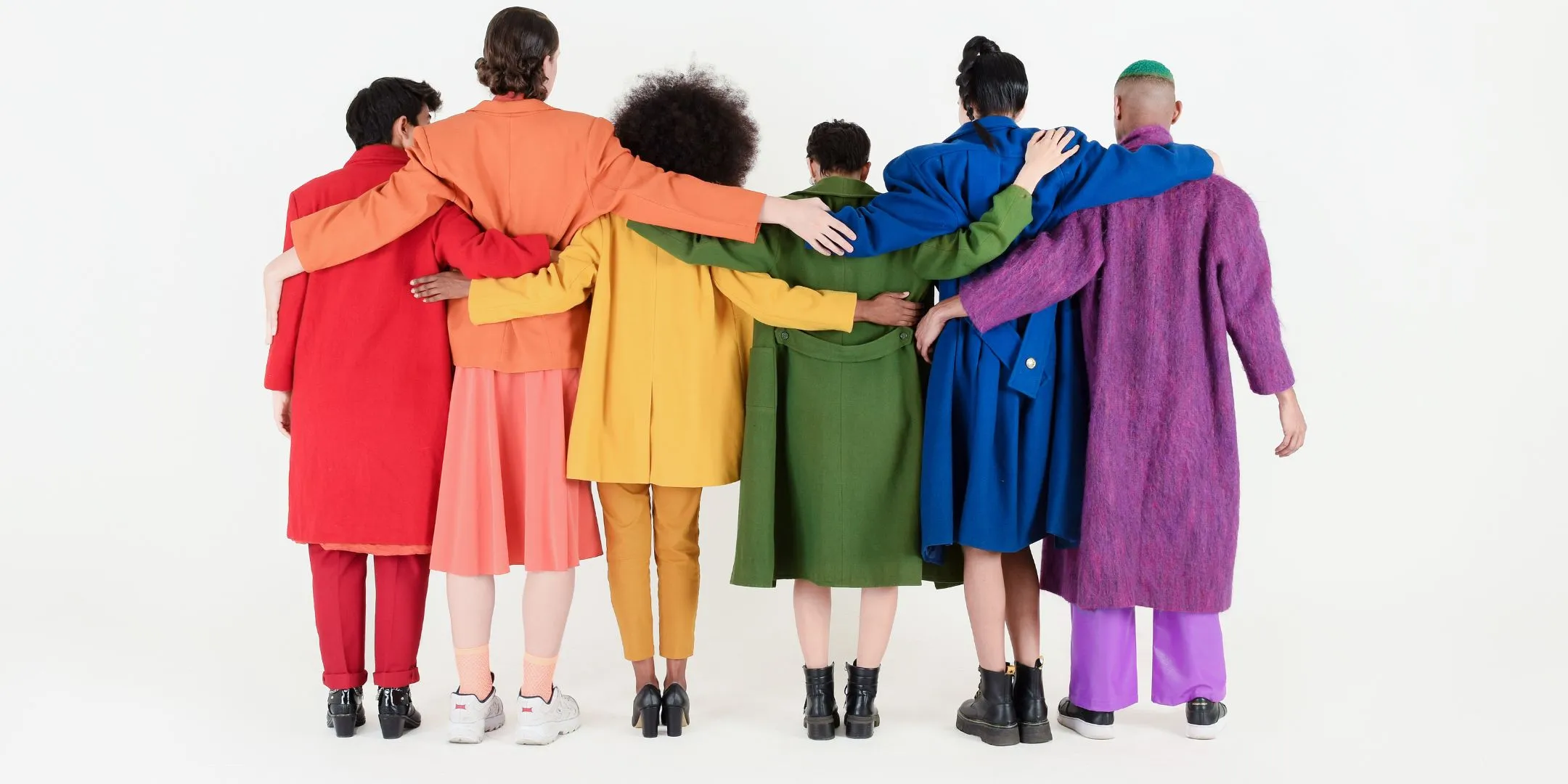Diverse group of six adults standing with their backs to the camera, arms around one another, wearing brightly coloured clothing against a white background.