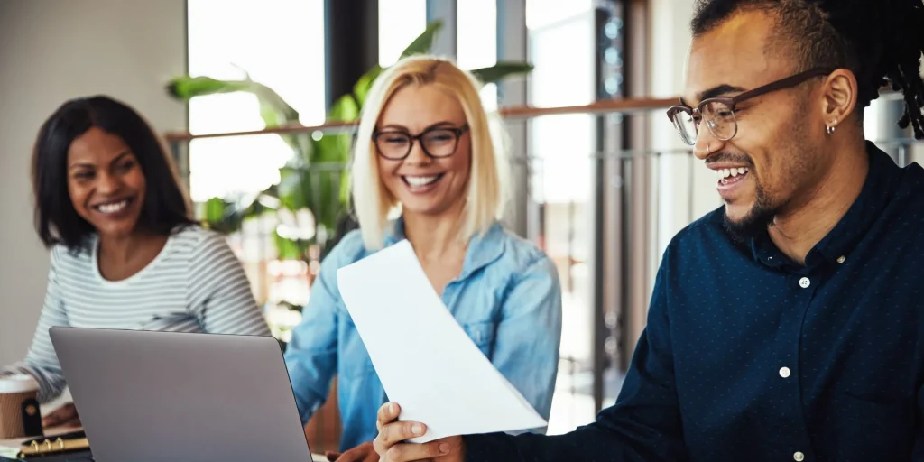 Three colleagues smile while reviewing a document together at a table with a laptop, collaborating in a bright office setting.