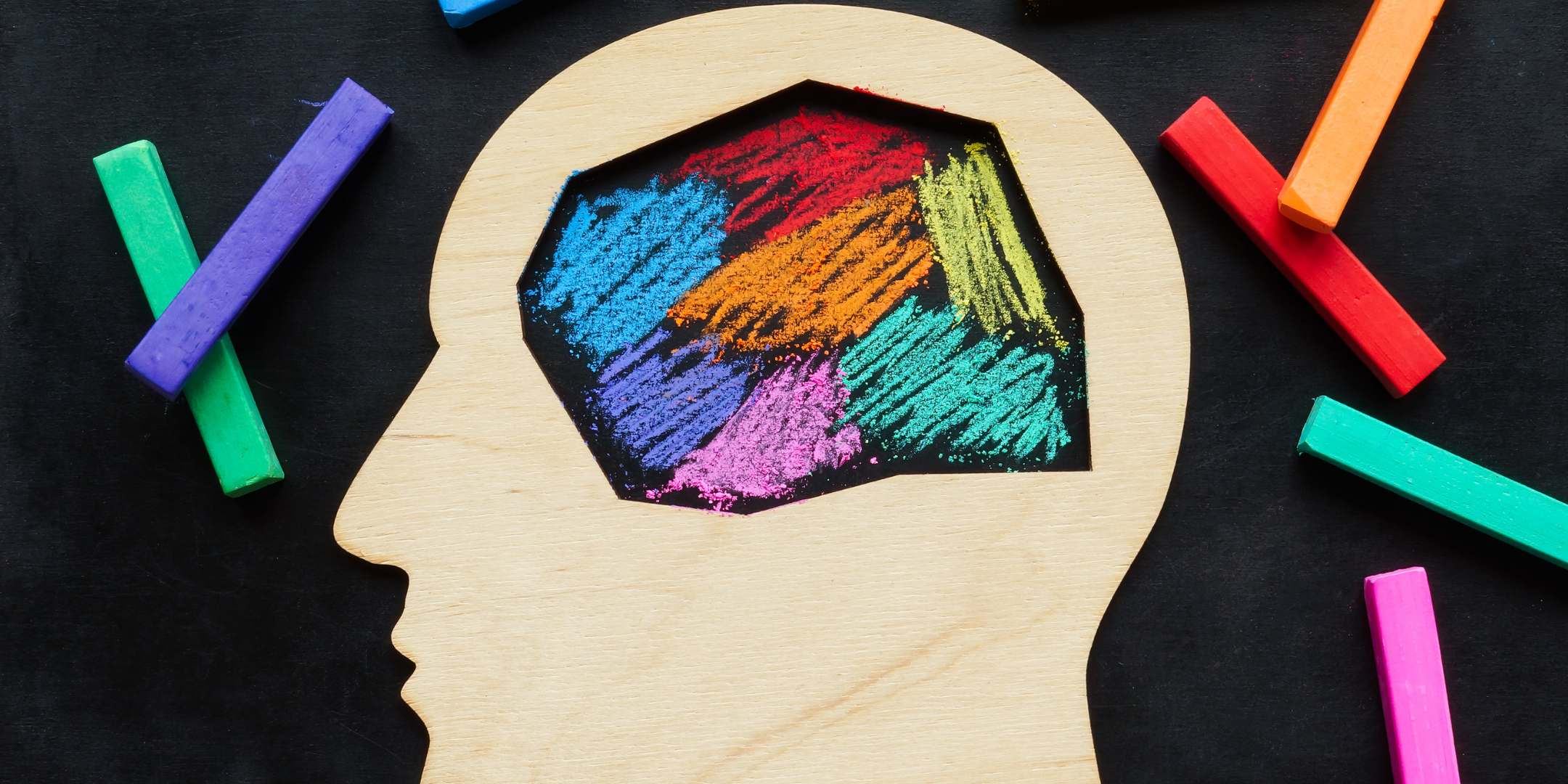 A wooden silhouette of a human head with an open brain area filled with brightly coloured chalk strokes, surrounded by colourful chalk pieces on a dark background.
