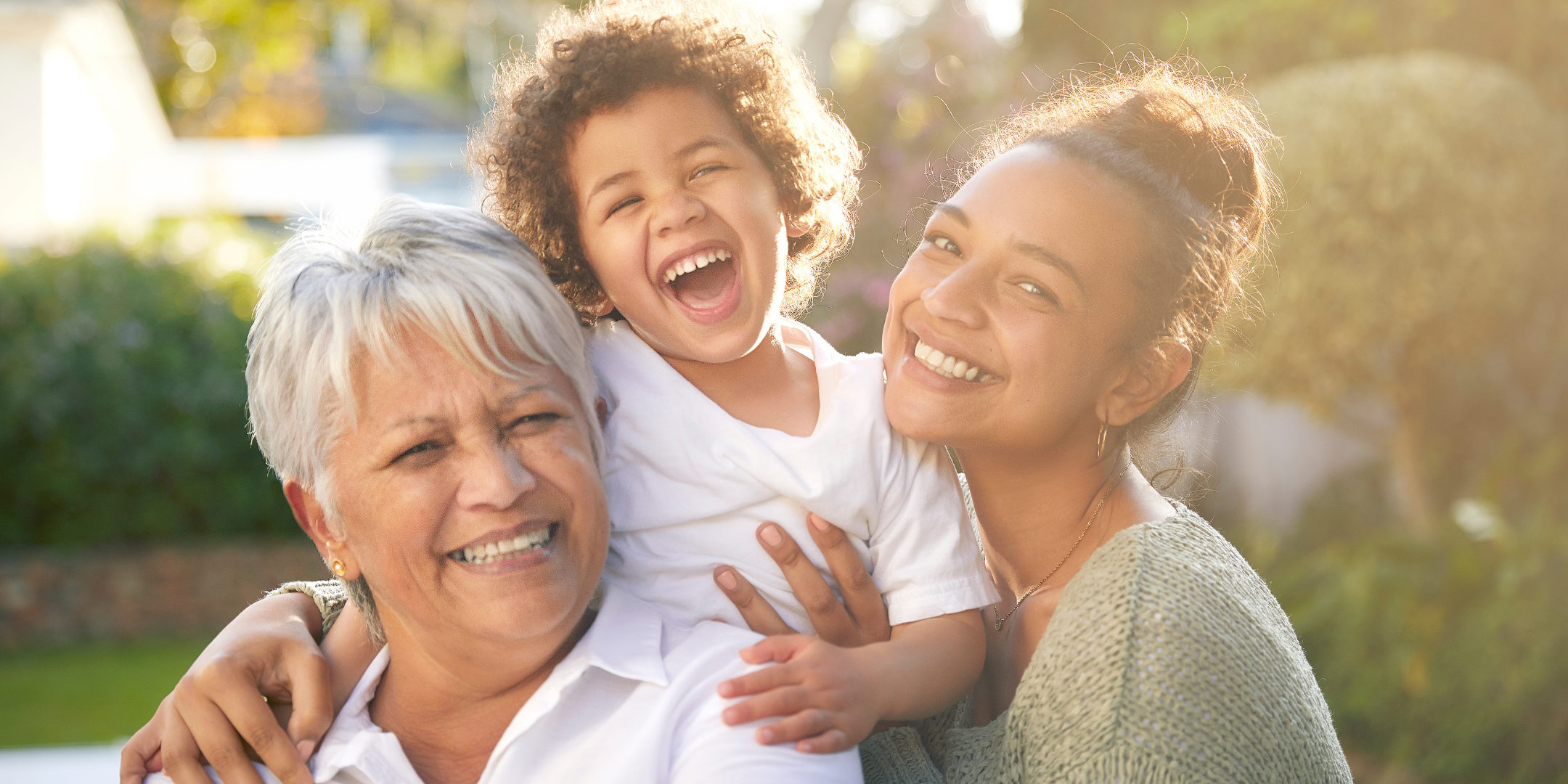 Three generations of a family smile and laugh together outdoors, with a young child held between two women in warm sunlight.