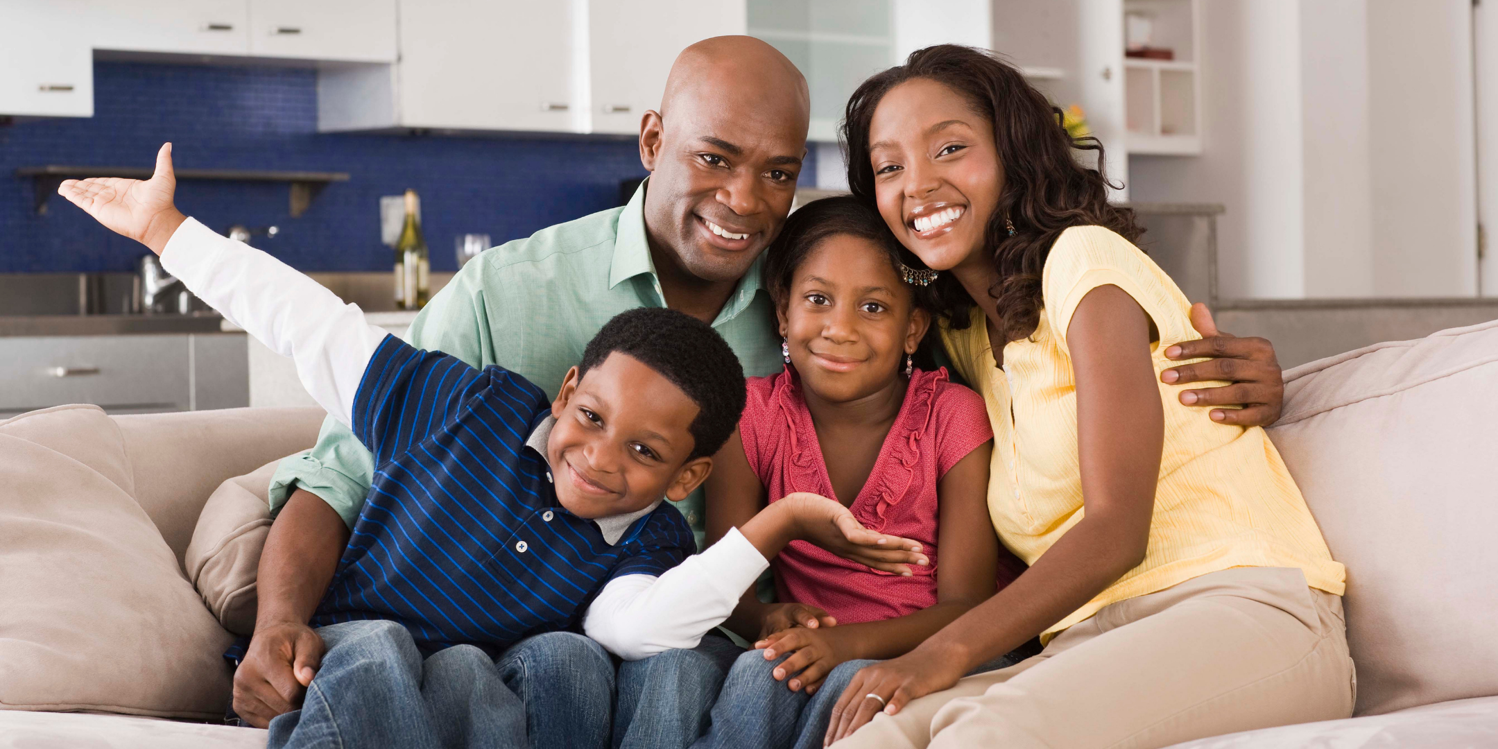 A smiling family of four sits closely together on a sofa at home, with two parents and two children sharing a warm, playful moment.