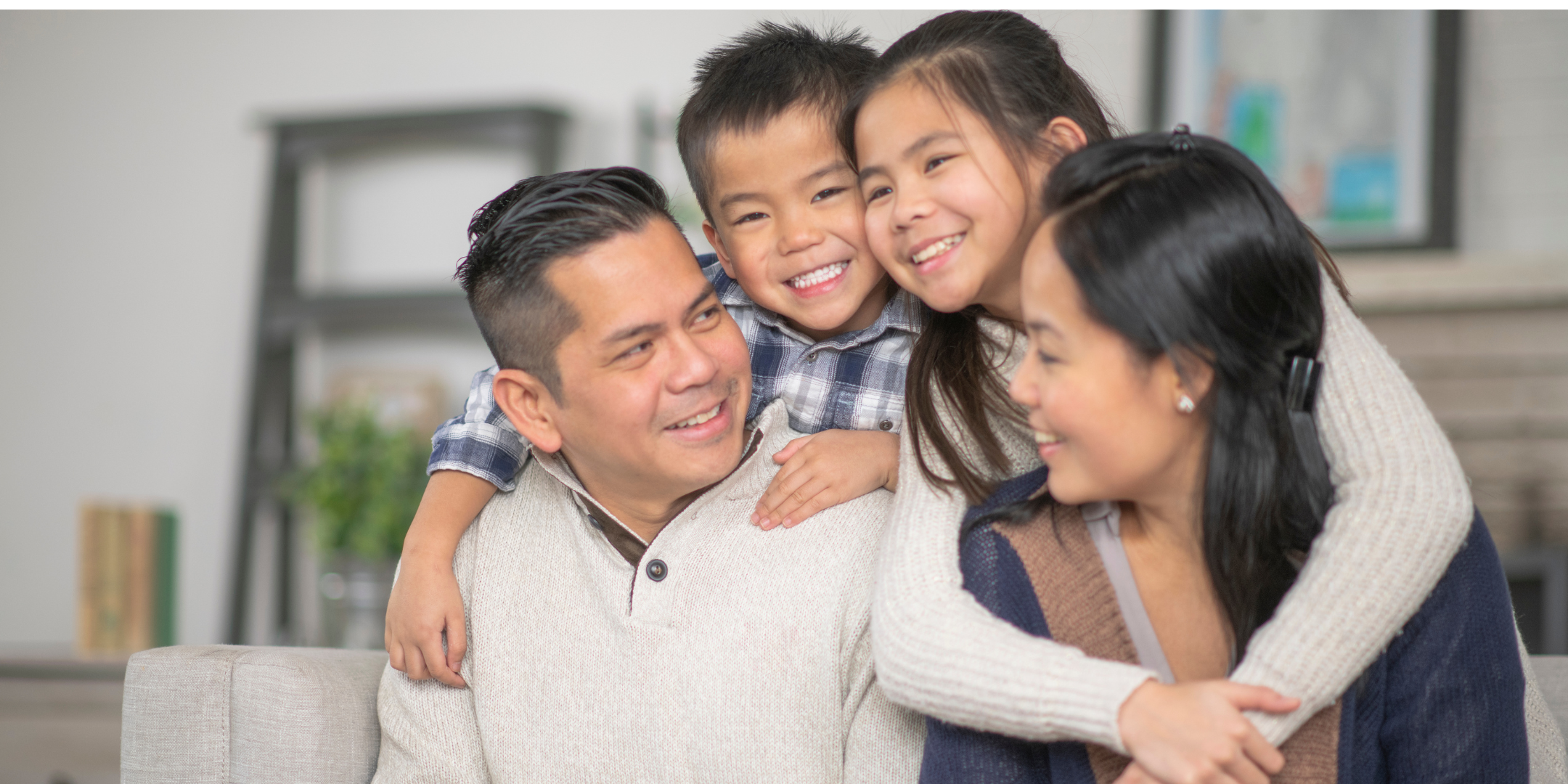 A smiling family of four sits closely together on a sofa, with two children affectionately hugging their parents.