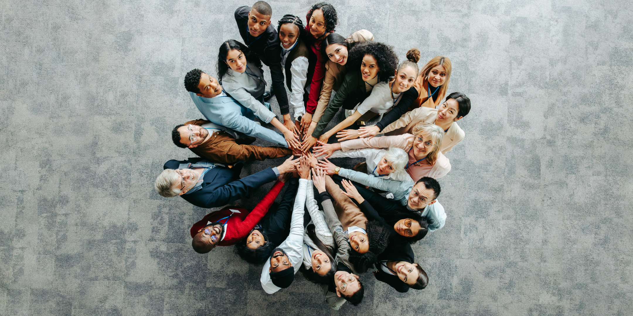Overhead view of a diverse, multigenerational group gathered in a circle with their hands joined together in the centre, representing community and collaboration.